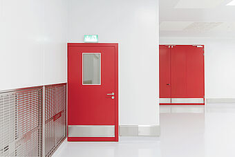 Turnkey hygiene room/clean room from Plattenhardt+Wirth GmbH. A stainless steel lock with directions serves as access control in a white-tiled hallway. All connections are hygienic and sterile. The door is red next to a stainless steel ventilation grille.