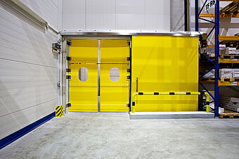 Gelbe Pendeltüre mit vorgesetztem gelben Brandschutztor/Kuehlraumtuer in einem Kühlraum zur Sicherstellung des Brandabschnittes. Ein markierter Anfahrschutz sichert das nebenstehende Hochregal vor Schäden.